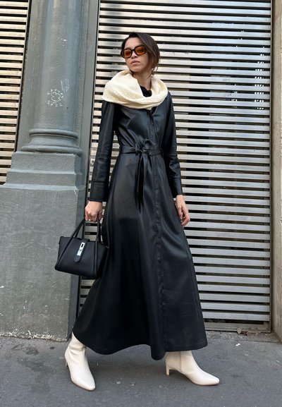 Abrigo de cuero negro con correa en la cintura, combinado con una bufanda de punto color crema, un bolso negro estructurado y botines blancos de punta. 