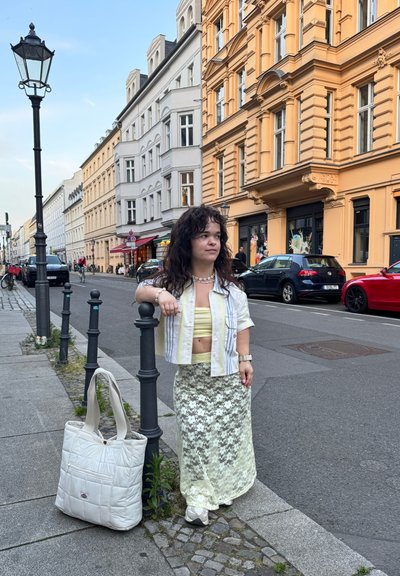 Bolso tote blanco acolchado con asas largas, camisa de rayas de manga corta sobre un top corto amarillo y una falda verde de encaje. Fondo de calle de la ciudad.