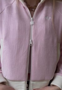 Personne portant une veste zippée côtelée rose clair avec des poignets blancs et le logo Adidas, visible du cou à la taille avec des colliers en or superposés.