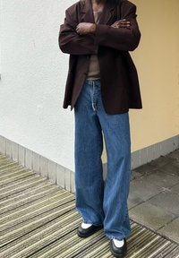 Brūns žakets virs bēša krekla, brīvi zili džinsi un melnas kurpes ar baltām akcentiem. Izteikti tekstūras un brīva silueta.
