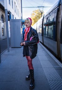 Svart skinnjakke over en svart kjole, røde strømpebukser med en tatovering, rødt og rosa strikket hodeplagg, kraftige svarte snørestøvler.