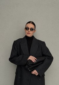Manteau grande taille boutonné gris foncé avec larges revers, col roulé noir, lunettes de soleil rondes foncées, et une pochette en cuir noire pliée.