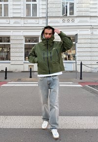 Zelena vodoodporna jakna s kapuco, z zadrgo spredaj in dvema črnima žepoma na prsih, v kombinaciji s svetlo modrimi džinsi in športnimi copati s srebrnimi detajli.