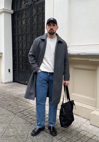 Cappotto lungo grigio sopra un maglione bianco, jeans in denim blu, sneaker nere e una borsa nera. Cappello piatto completa l'outfit. Ambientazione esterna lastricata.