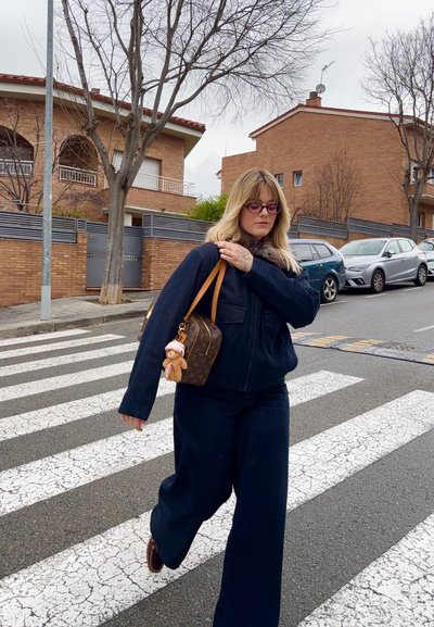 Mujer de cabello rubio y gafas moradas vestida con ropa oscura cruza una calle por un paso de peatones, llevando un bolso marrón con un llavero de oso de peluche.
