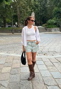 Ελαφρύ λευκό πουκάμισο πάνω από μια εφαρμοστή λευκή αμάνικη μπλούζα, συνδυασμένο με τζιν σορτς. Καφέ ψηλές μπότες και μαύρη τσάντα. Αστικός χώρος με πέτρινο πλακόστρωτο.