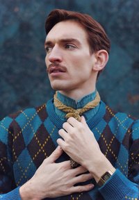 Homme à moustache ajustant un foulard beige, portant un pull bleu à motif losanges, une montre digitale et une bague à motifs, regardant vers la gauche sur un fond sombre.