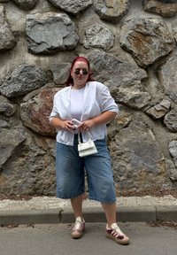 Casaco branco, leve e de manga curta sobre uma blusa branca; culotes de ganga azul-claro; sapatos rosa e castanhos; pequena mala branca. Fundo pedregoso.