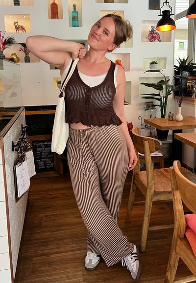 Chaleco corto de croché marrón sobre una camiseta blanca, combinado con pantalones anchos a rayas marrones. Modela un bolso tote y lleva zapatos blancos con detalles grises.