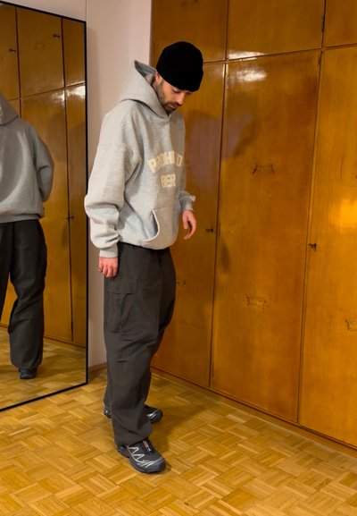 Sudadera con capucha gris con el texto "PROHIBIDO BEBER", pantalones cargo negros y zapatillas deportivas grises; de pie cerca de un espejo y un armario de madera, en un entorno interior.