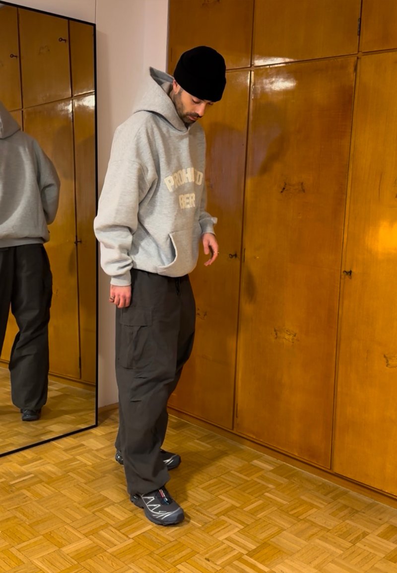 Sweat à capuche gris avec le texte « PROHIBIDO BEBER », pantalons cargo noirs et chaussures de sport grises ; debout près d'un miroir et d'une armoire en bois, en intérieur.