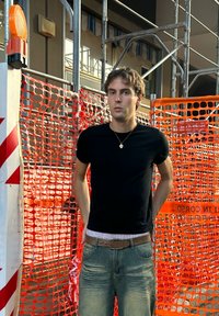 T-shirt nera aderente, jeans azzurri con tasche, in piedi vicino a barriere di costruzione arancioni e impalcature sullo sfondo.