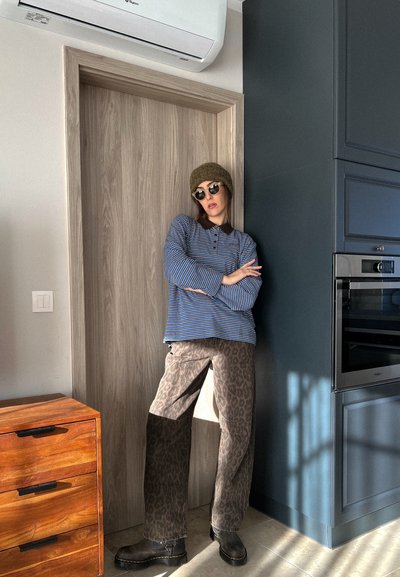 Camisa de manga larga a rayas azul y blanca, pantalones de leopardo marrón, botas marrones y gorro de punto verde, de pie junto a una puerta de madera y mobiliario moderno.