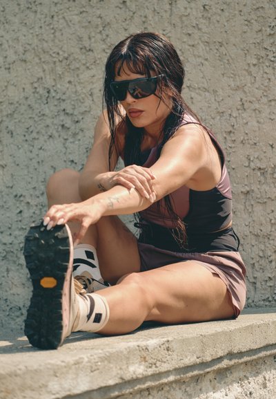 Conjunto deportivo que incluye una camiseta negra ajustada y pantalones cortos morados. Gafas de sol negras. Zapatillas de color marrón claro con suelas texturizadas y detalles en blanco.