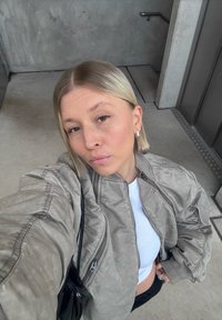 Lichtgrijze bomberjack met textuur en geribde manchetten, gedragen over een witte geribde top. Zwarte handtas zichtbaar. Casual setting.