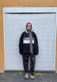 Jeune femme portant un hijab gris, un pull noir et gris, un pantalon à motifs et des baskets, debout sur un sol carrelé avec des feuilles éparpillées.