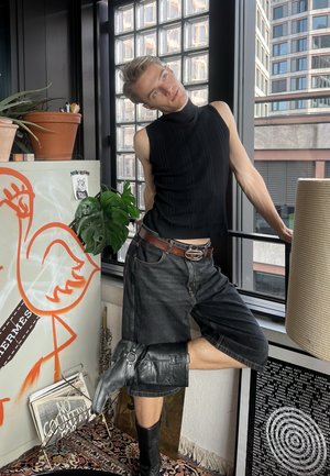 Persona con camiseta negra sin mangas y pantalones cortos largos posando junto a la ventana, apoyándose hacia atrás con una pierna levantada, rodeada de plantas y obras de arte.