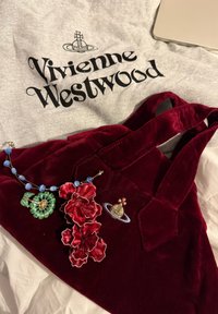 Rød fløyelsveske med to håndtak, utsmykket med blomstermotiver og et rom-tema merke. Grå skjorte med "Vivienne Westwood" i svart.