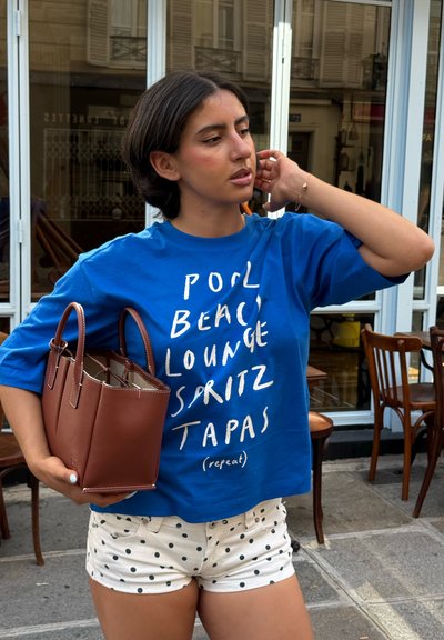 Joven mujer con camiseta azul con gráfico y shorts blancos con lunares sostiene un bolso marrón, de pie fuera de una cafetería con sillas y mesas de madera.