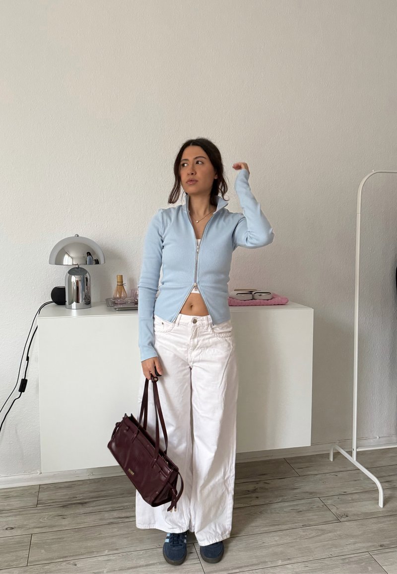 Žena v svetlomodrom zipsovom vrchu, bielych širokých nohaviciach, držiaca tmavohnedú kabelku, stojaca v minimalistickej miestnosti s drevenou podlahou a bielymi stenami.