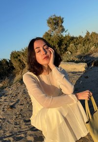 Rochie lungă, semi-transparentă, de culoare bej deschis, cu fustă fluidă. Subiectul ține o geantă verde și stă pe nisip, având verdeață în fundal.