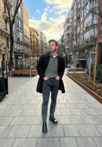 Uomo in un cappotto nero sopra una polo verde e jeans neri, con stivali neri, che sta in una strada pavimentata fiancheggiata da alberi e edifici.