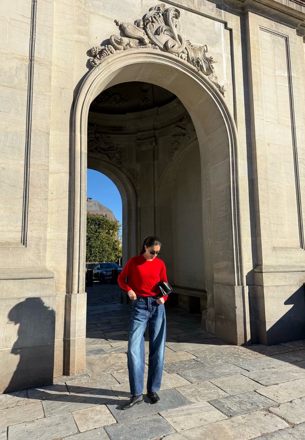 Una mujer con un jersey rojo y vaqueros está de pie bajo un arco de piedra ornamentado, sosteniendo un bolso negro a plena luz del sol.