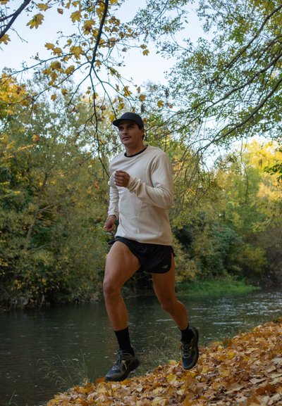 Un corredor masculino con una camiseta de manga larga beige claro y pantalones cortos negros trota a lo largo de una orilla de río bordeada de hojas de otoño, vistiendo una gorra negra y zapatillas para correr.