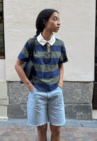 Persona con cabello oscuro y rizado, vistiendo una camisa a rayas verde y azul marino con cuello blanco, pantalones cortos de mezclilla y un bolso negro al hombro, de pie sobre un pavimento de piedra.