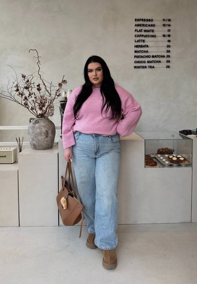 Suéter rosa, jeans celestes, botas marrones, sosteniendo un bolso marrón. Jarrón gris con ramas y algodón al lado de una exhibición de productos horneados.