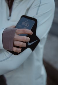 Brassard de smartphone noir avec un logo Nike, fixé solidement sur la manche d'une veste blanche, affichant une horloge numérique sur le téléphone.
