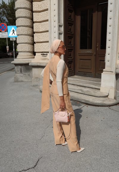 Conjunto de dos piezas beige con chaleco ajustado y pantalones de pierna ancha, combinado con un bolso rosa. Tejido suave, diseño minimalista y tonos pastel claros.