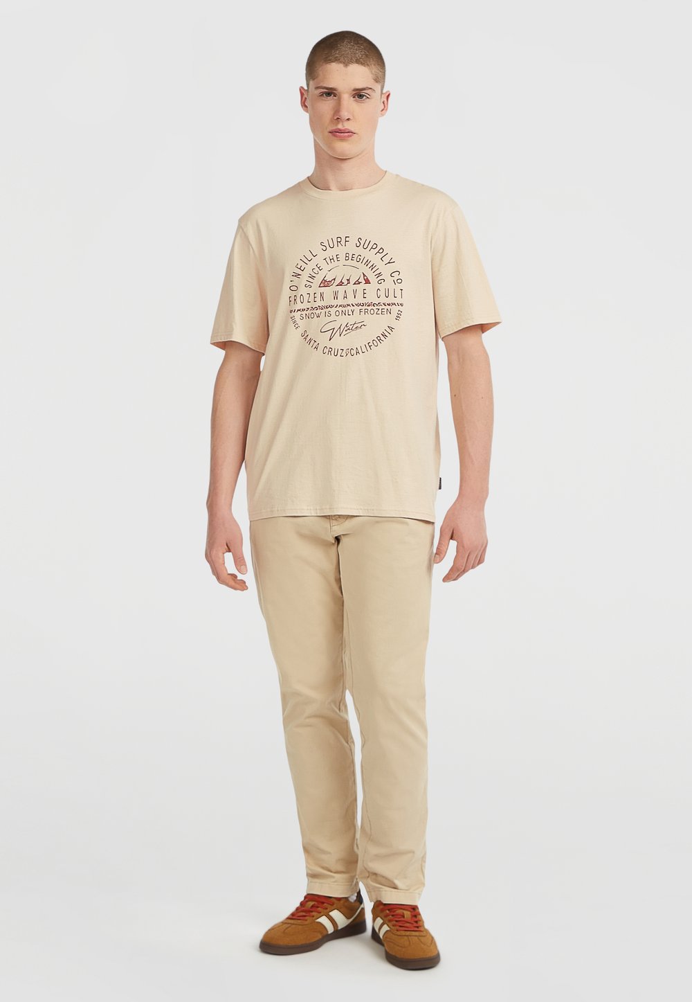 Beige bomulds t-shirt med cirkulær rød grafisk tekst; parret med beige bukser og brune sneakers med hvide striber og røde snørebånd.