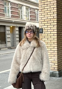 Kvinde iført briller, leopardprint-hat, beige kunstpelsjakke og brune bukser med striber, stående på en bygade nær en murstenssøjle.