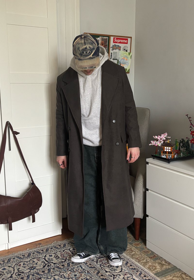 Personne portant un long manteau sombre, un sweat à capuche gris, un jeans large noir, des baskets et une casquette de baseball usée, se tenant dans une pièce avec des meubles et des œuvres murales.