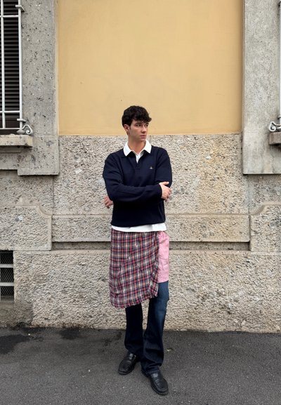 Joven de pie con los brazos cruzados, vistiendo un suéter azul marino, camisa blanca con cuello, falda de cuadros y rosa superpuesta sobre jeans oscuros, junto a una pared de piedra.