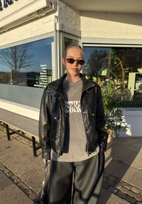 Veste en cuir noire avec ferrures argentées, t-shirt gris avec texte blanc, pantalon noir à jambes larges, et sac à main noir avec une bandoulière chaîne argentée.