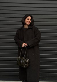 Manteau en laine marron avec de gros boutons, modèle surdimensionné et large col ; assorti à un sac à main sombre et brillant et une bague argentée à la main.