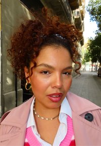 Zbliżenie osoby o kręconych, rudawych włosach, ubrane w różowy płaszcz, białą koszulę i perłowy naszyjnik w miejskim otoczeniu.