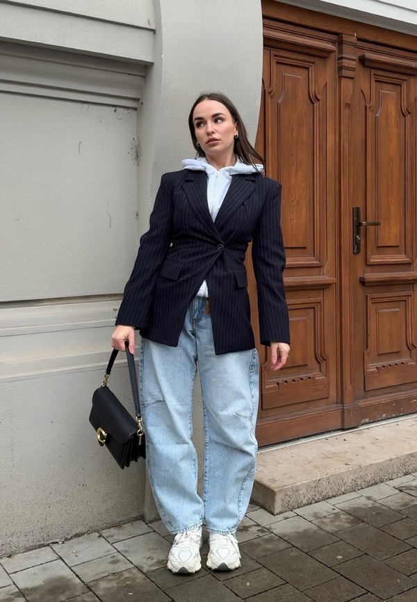 Kvinne i en mørkeblå dressjakke med striper over en hvit hettegenser, lyseblå jeans, hvite joggesko, holder en svart håndveske, står ved en tredør.