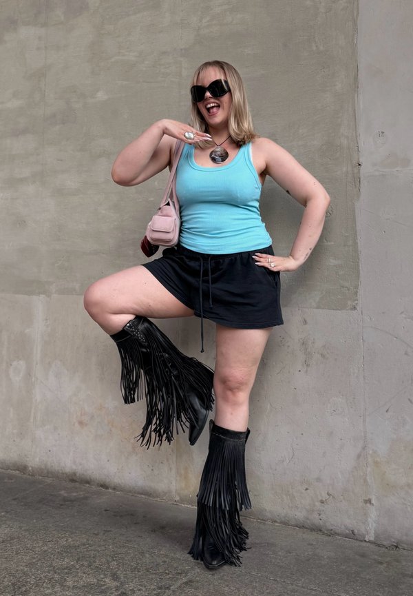 Persoon draagt een blauw tanktopje, zwarte korte broek en zwarte laarzen met franjes, poseert met één been omhoog en lacht tegen een effen muur.