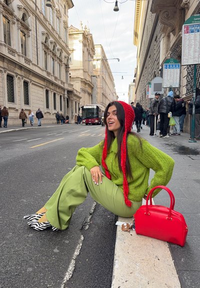 Suéter verde brillante de gran tamaño, pantalones verdes de pierna ancha, zapatos con estampado de cebra y un bolso rojo. Escenario al aire libre con edificios al fondo.