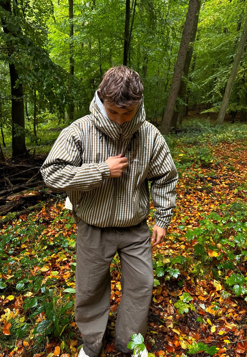 Jeune personne portant une veste rayée à capuche, debout sur un sentier forestier recouvert de feuilles, entourée d'arbres verts en automne.