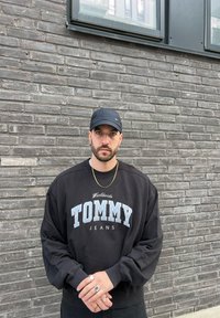 Fekete pulóver "TOMMY JEANS" kék és fehér felirattal. Fekete sapkával és több gyűrűvel viselve, sz�ürke téglafal előtt.
