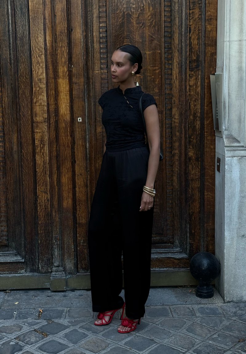 Femme vêtue d'une tenue noire et de sandales rouges à talons hauts, debout devant une grande porte en bois sur un pavé en pierre.
