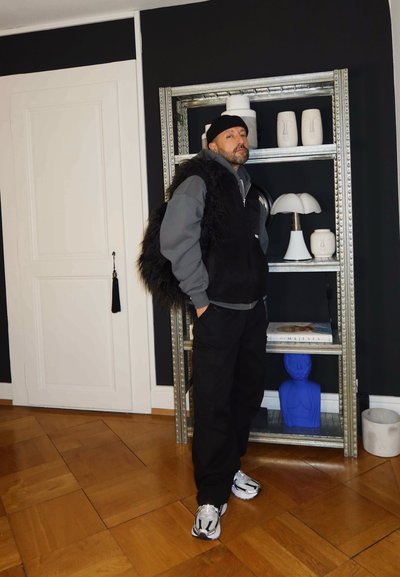 Hombre con un gorro negro, sudadera con capucha, chaleco de piel y zapatillas de deporte se encuentra de pie frente a una estantería de metal con esculturas modernas en blanco y azul.