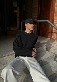 Črni pleteni pulover z razcefranimi robovi, kombiniran z ohlapnimi belimi hlačami. Oseba nosi črno kapo in sedi na kamnitih stopnicah.