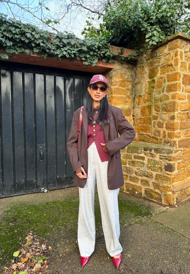 Femme portant une casquette bordeaux, des lunettes de soleil, un blazer, un cardigan boutonné, un pantalon large gris et des chaussures rouges pointues, debout près d'un mur en pierre et d'une porte noire.
