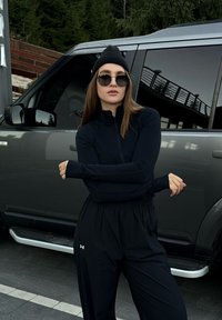 Conjunto atlético preto com uma blusa de mangas longas com fecho e calças folgadas. Os acess�órios incluem um gorro preto e óculos de sol. Veículo cinza ao fundo.