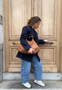 Femme en blazer bleu marine et jean tenant un sac à main beige tout en ouvrant une grande porte en bois avec une poignée en laiton.